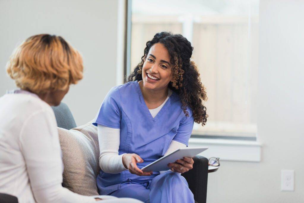 The mid adult female visiting nurse chats with the unrecognizable senior adult female patient. The nurse enters patient responses on the digital tablet.